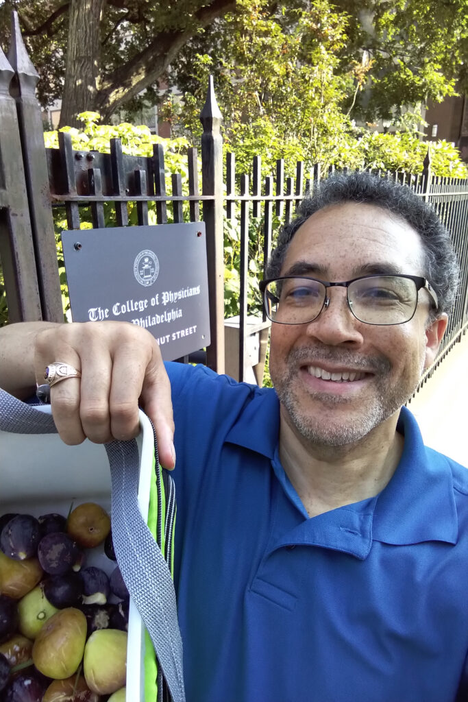 A man smiles while holding plums and a booklet outdoors.